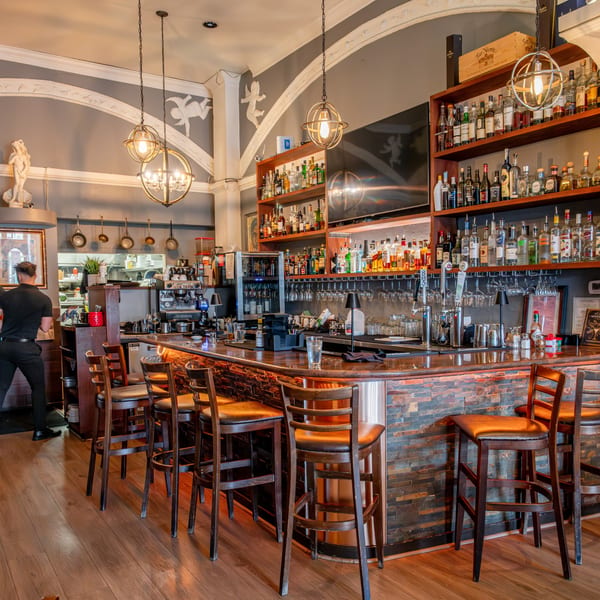 A bartender works behind a fully stocked bar with wooden stools and decorative lighting at Roma Restaurant.