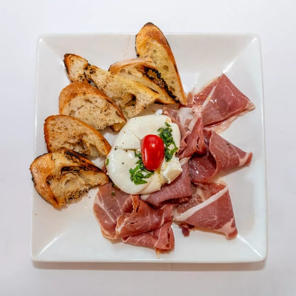 A white plate with burrata cheese, prosciutto, and toasted bread, garnished with a cherry tomato and herbs.