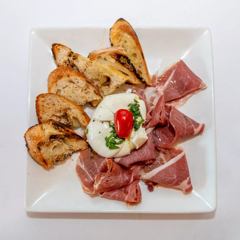 A white plate with burrata cheese, prosciutto, and toasted bread, garnished with a cherry tomato and herbs.