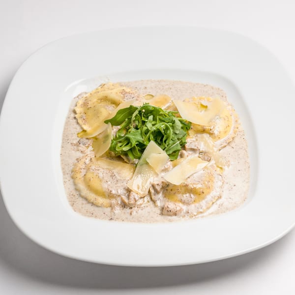 Ravioli in a creamy mushroom sauce, topped with fresh arugula and parmesan shavings on a white plate.