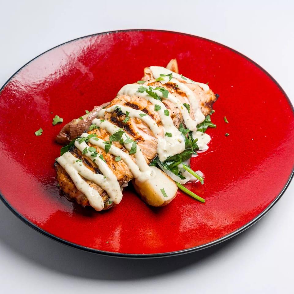 Grilled chicken with creamy sauce and fresh herbs, served on a bed of greens on a red plate.